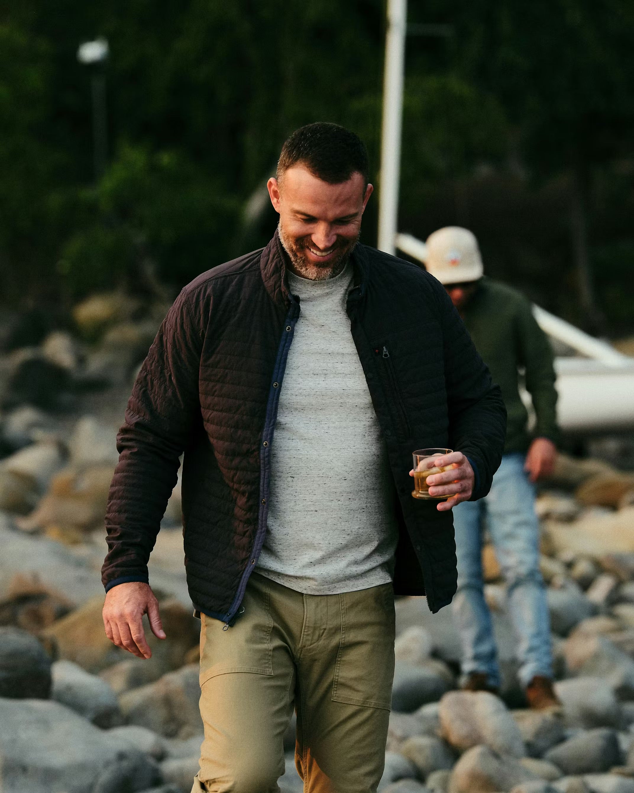 Man walking outdoors on a rocky path holding a drink, with another person in the background.