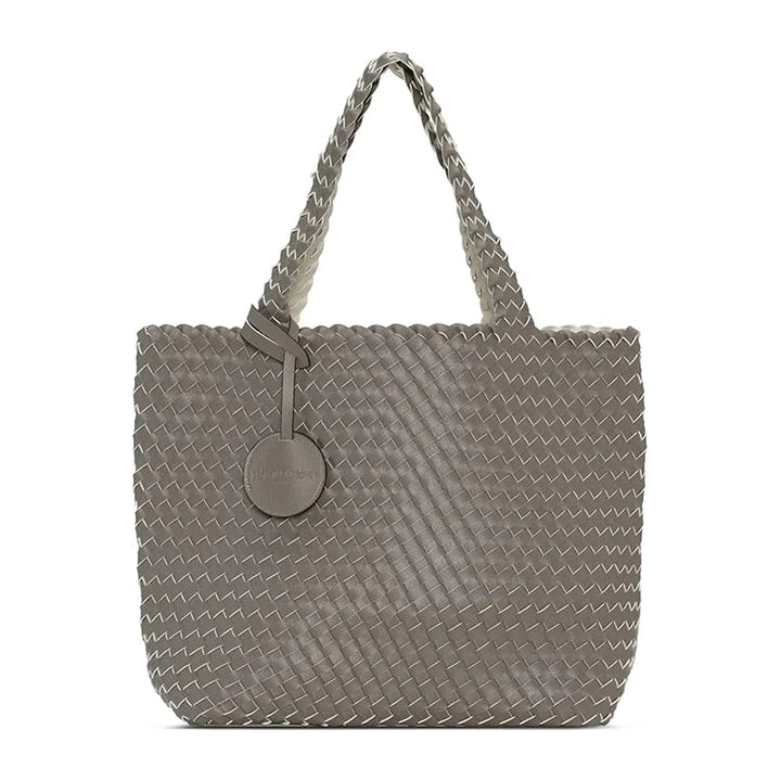 A woven leather tote bag in a taupe-gray color, featuring braided shoulder straps and a circular leather brand tag on the front, set against a plain white background.