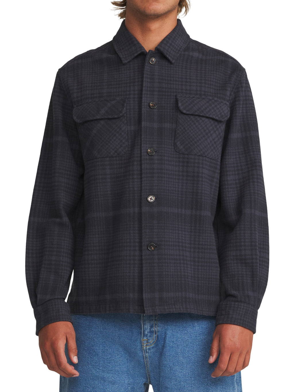 A man wearing a dark charcoal gray and black plaid button-up shirt jacket with two chest flap pockets, paired with blue jeans, against a plain white background.