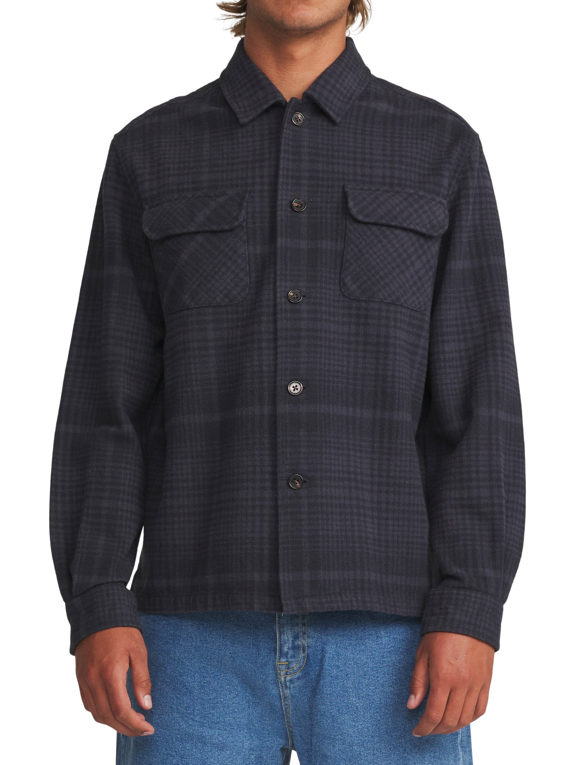 A man wearing a dark charcoal gray and black plaid button-up shirt jacket with two chest flap pockets, paired with blue jeans, against a plain white background.