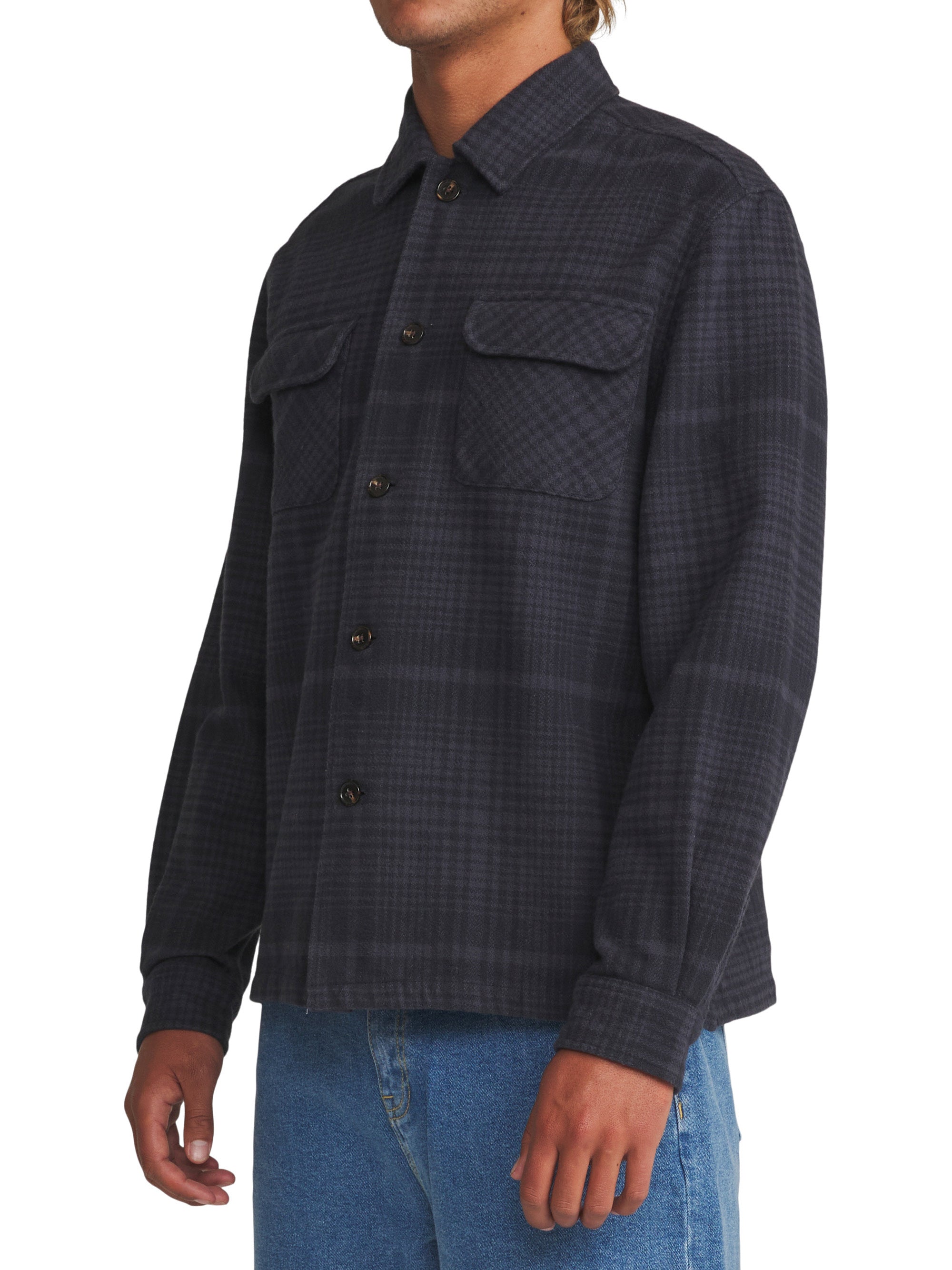 A man wearing a dark charcoal gray and black plaid button-up shirt jacket with two chest pockets, paired with blue jeans, shown from the waist up against a white background.