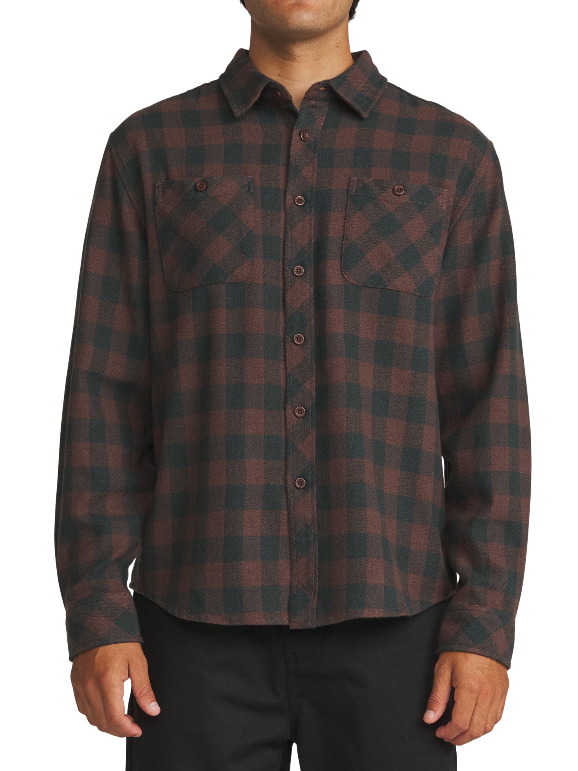 A man wearing a long-sleeve button-up flannel shirt with a dark green and brown plaid pattern, paired with black pants, against a plain white background.