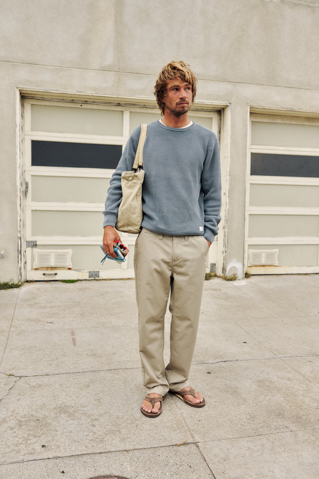 A man stands on a concrete sidewalk in front of a garage door, wearing a dusty blue crewneck sweatshirt, beige pants, and brown sandals, with a beige canvas shoulder bag slung over his shoulder.