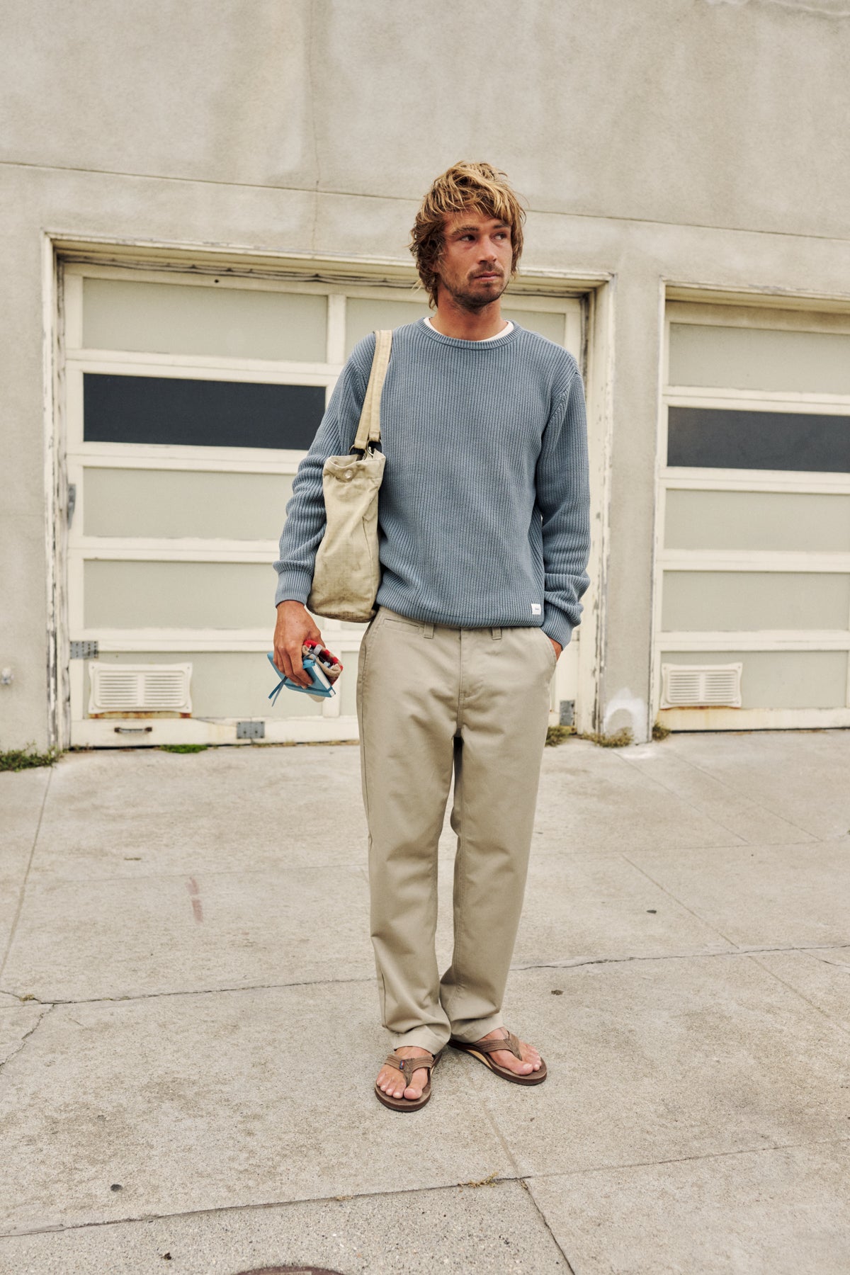 A man stands on a concrete sidewalk in front of a garage door, wearing a dusty blue crewneck sweatshirt, beige pants, and brown sandals, with a beige canvas shoulder bag slung over his shoulder.