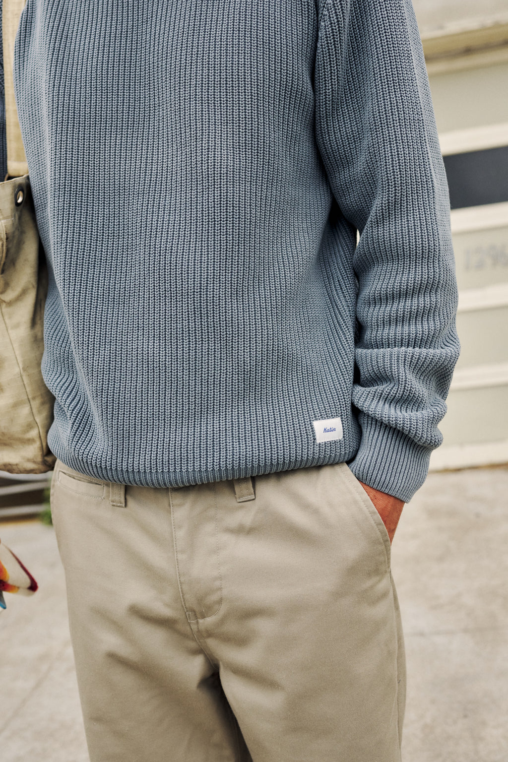 A close-up of a person wearing a ribbed knit sweater in a muted blue-gray color, paired with beige pants. The sweater has a small white rectangular brand tag near the lower hem.