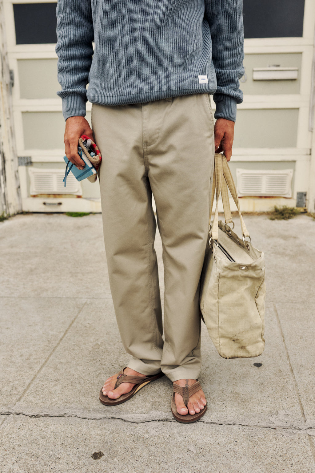 A person standing outdoors on a concrete surface wearing a ribbed blue-gray sweater, beige chinos, and brown sandals, holding a beige tote bag and a small blue item in one hand.