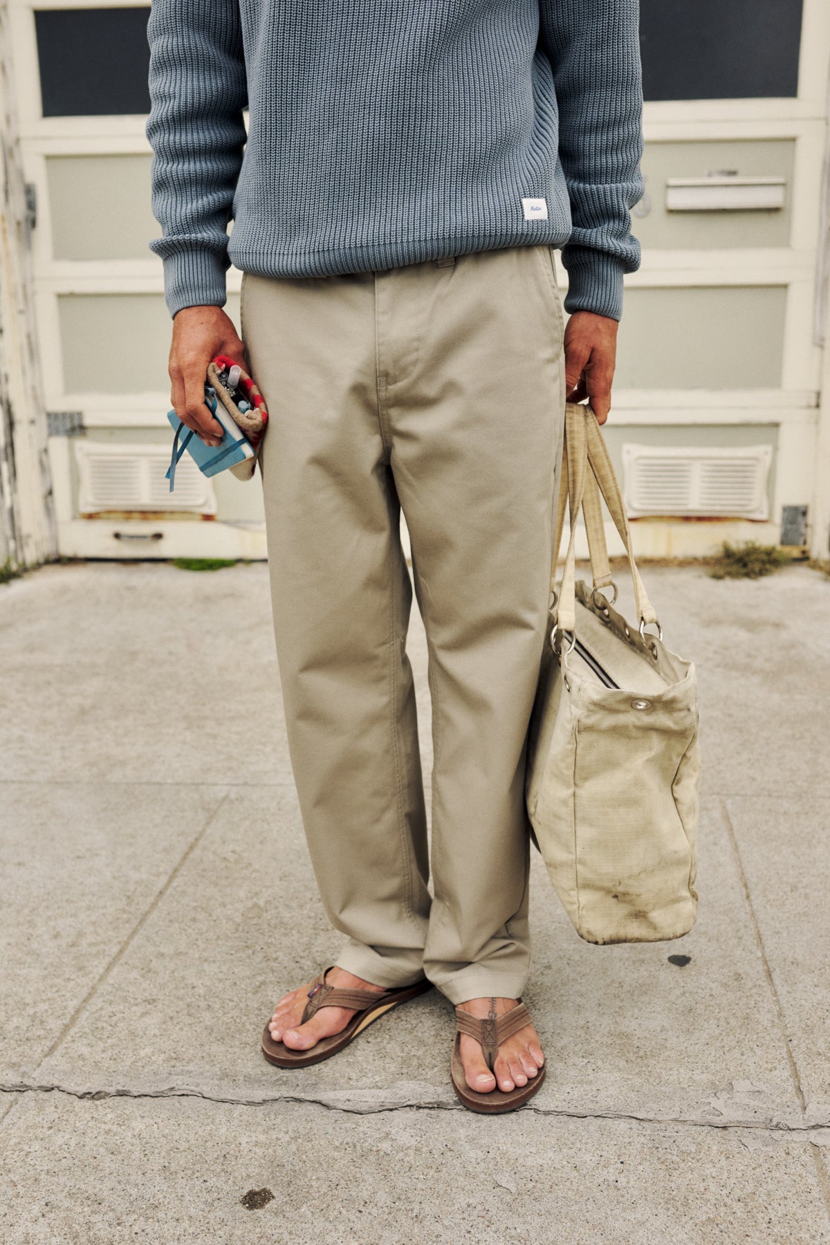 A person standing outdoors on a concrete surface wearing a ribbed blue-gray sweater, beige chinos, and brown sandals, holding a beige tote bag and a small blue item in one hand.
