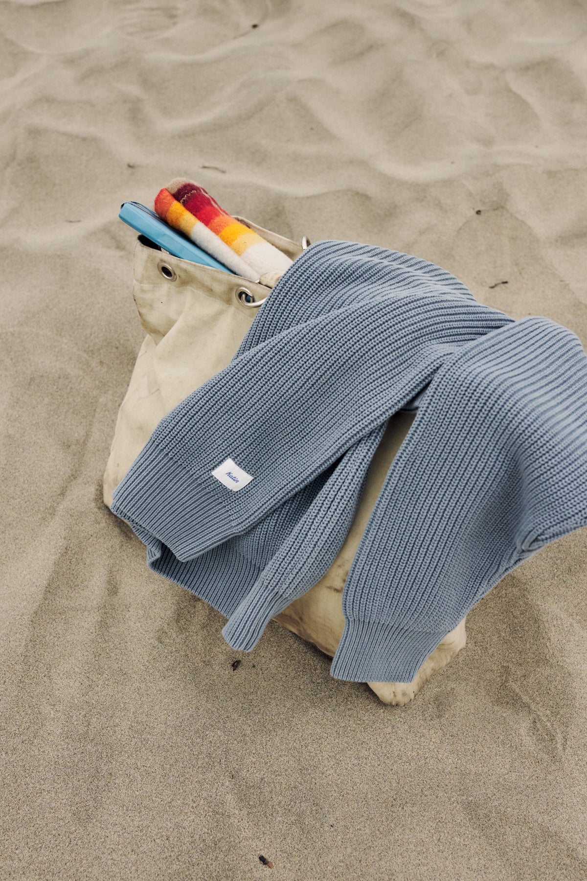 A light blue ribbed knit sweater draped over a beige canvas tote bag on a sandy beach, with colorful rolled towels peeking out from the top.