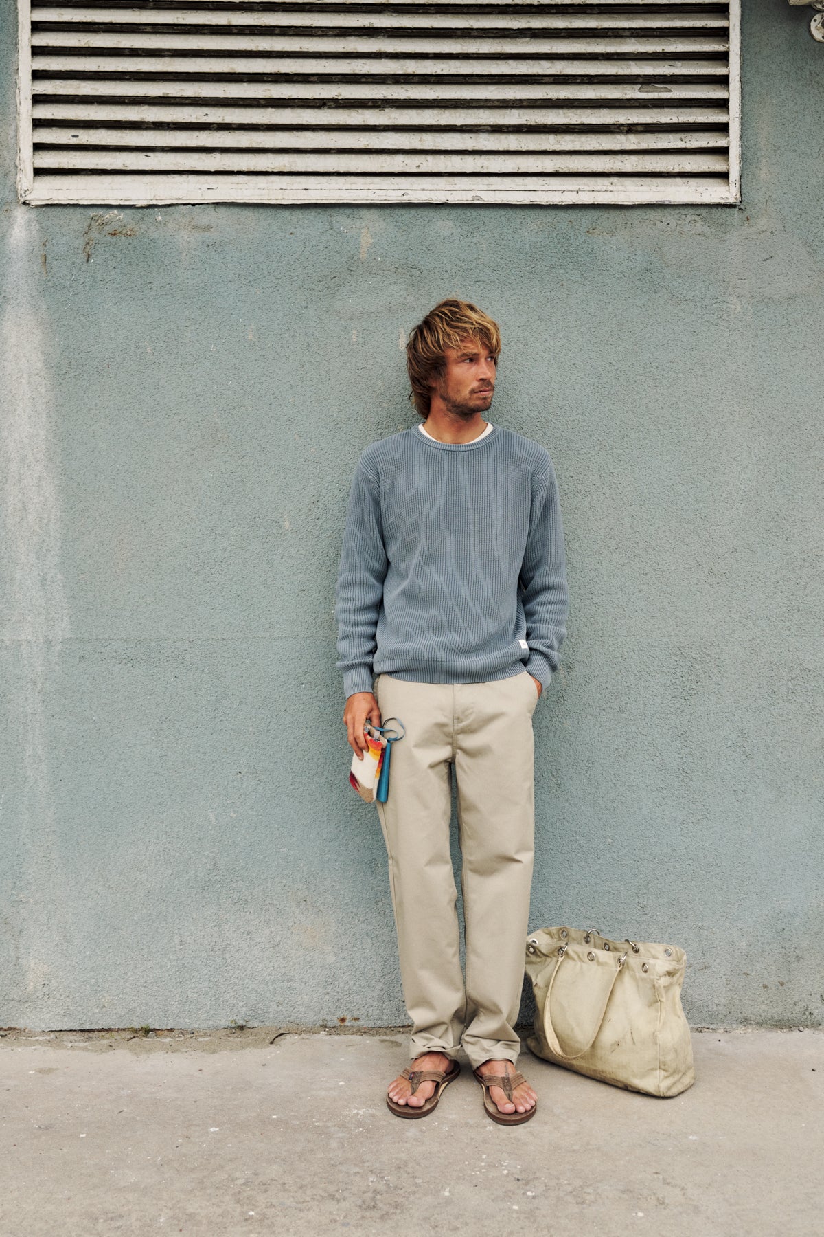 A man stands against a textured light blue-gray wall wearing a dusty blue-gray crewneck sweater, beige trousers, and brown sandals. A beige canvas tote bag rests on the concrete ground next to him.