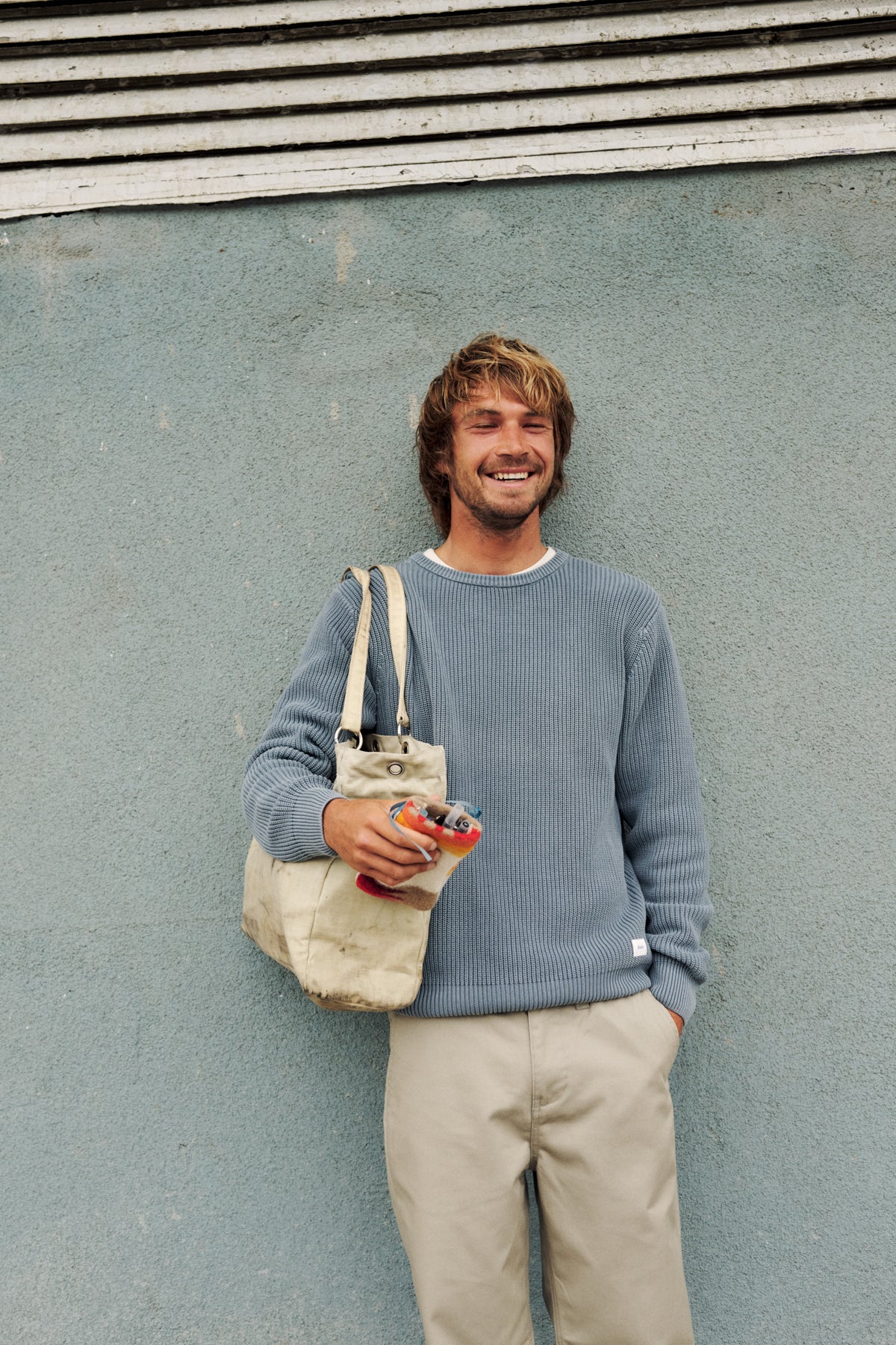 A smiling man with blond hair stands against a textured blue-gray wall, wearing a light blue ribbed knit sweater, beige pants, and carrying a beige canvas tote bag over his shoulder.