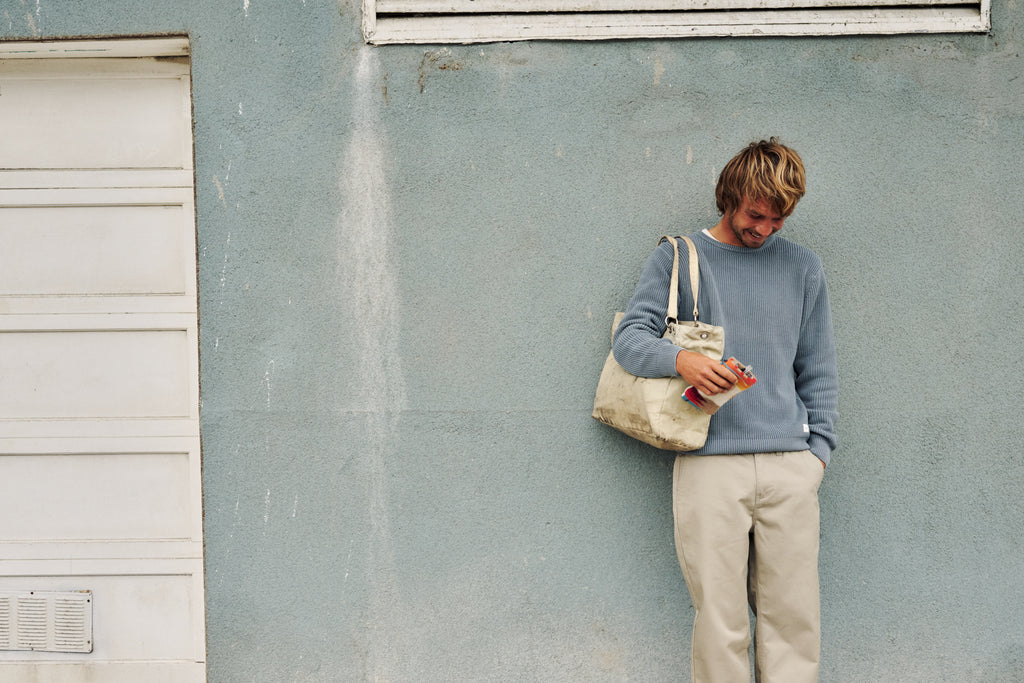A man with blond hair leans against a textured light blue-gray wall, wearing a dusty blue long-sleeve sweater and beige trousers. He is holding a cream canvas tote bag and a small red-and-white packaged item.