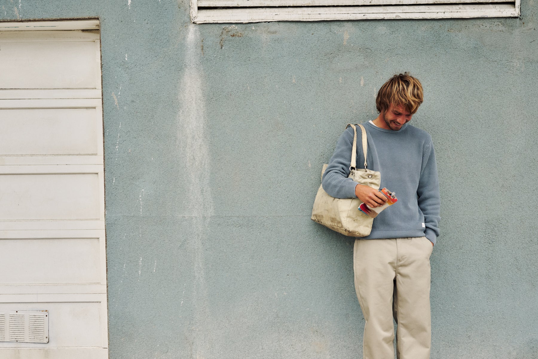 A man with blond hair leans against a textured light blue-gray wall, wearing a dusty blue long-sleeve sweater and beige trousers. He is holding a cream canvas tote bag and a small red-and-white packaged item.