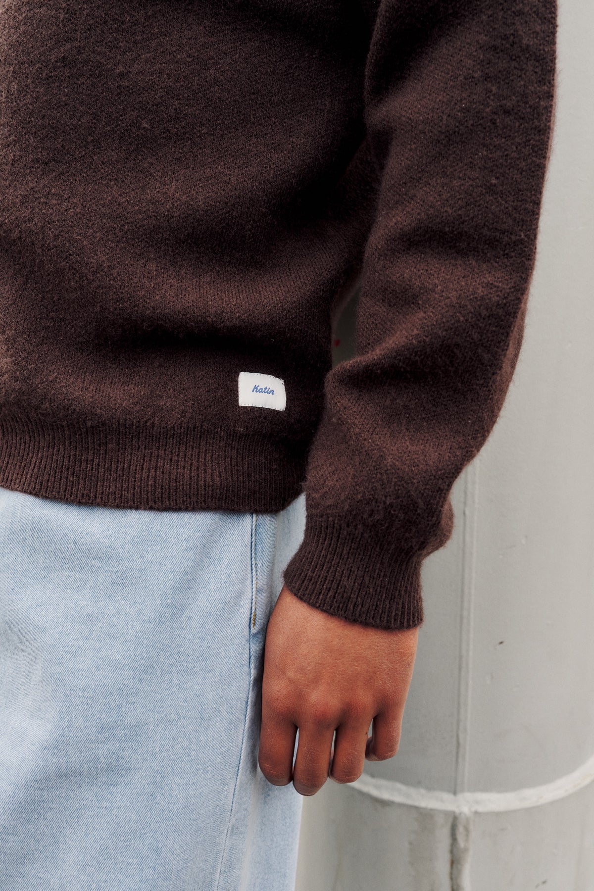 A close-up of a person wearing a dark brown knit sweater with a small white brand patch on the lower sleeve, paired with light blue jeans, standing against a light gray concrete wall.