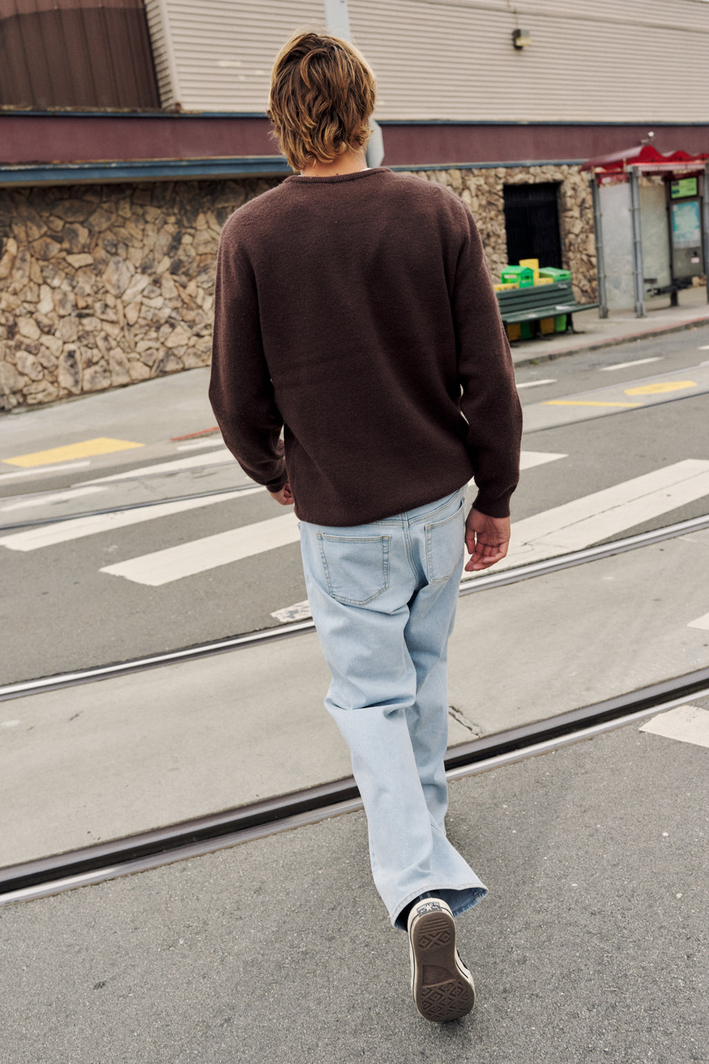A man walking away on a city street, wearing a dark brown long-sleeve knit sweater, light blue jeans, and dark sneakers.
