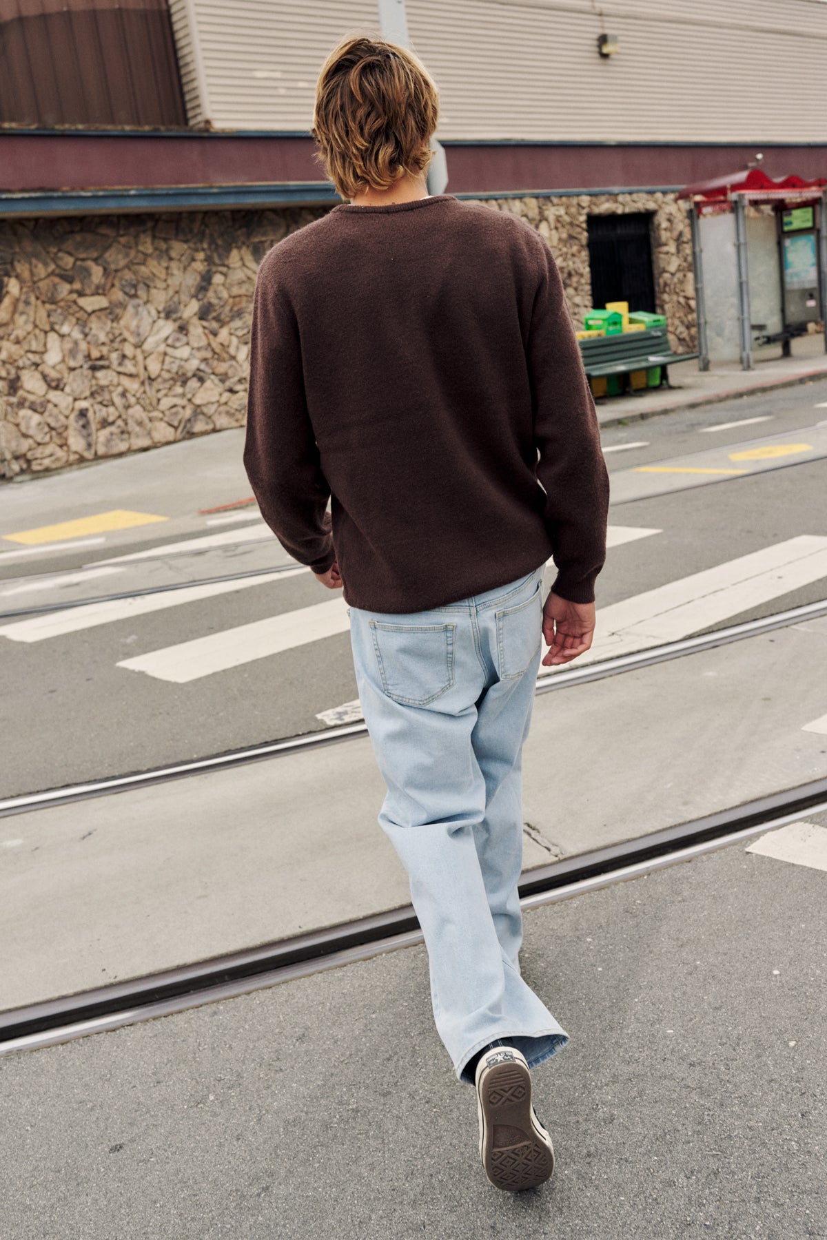 A man walking away on a city street, wearing a dark brown long-sleeve knit sweater, light blue jeans, and dark sneakers.