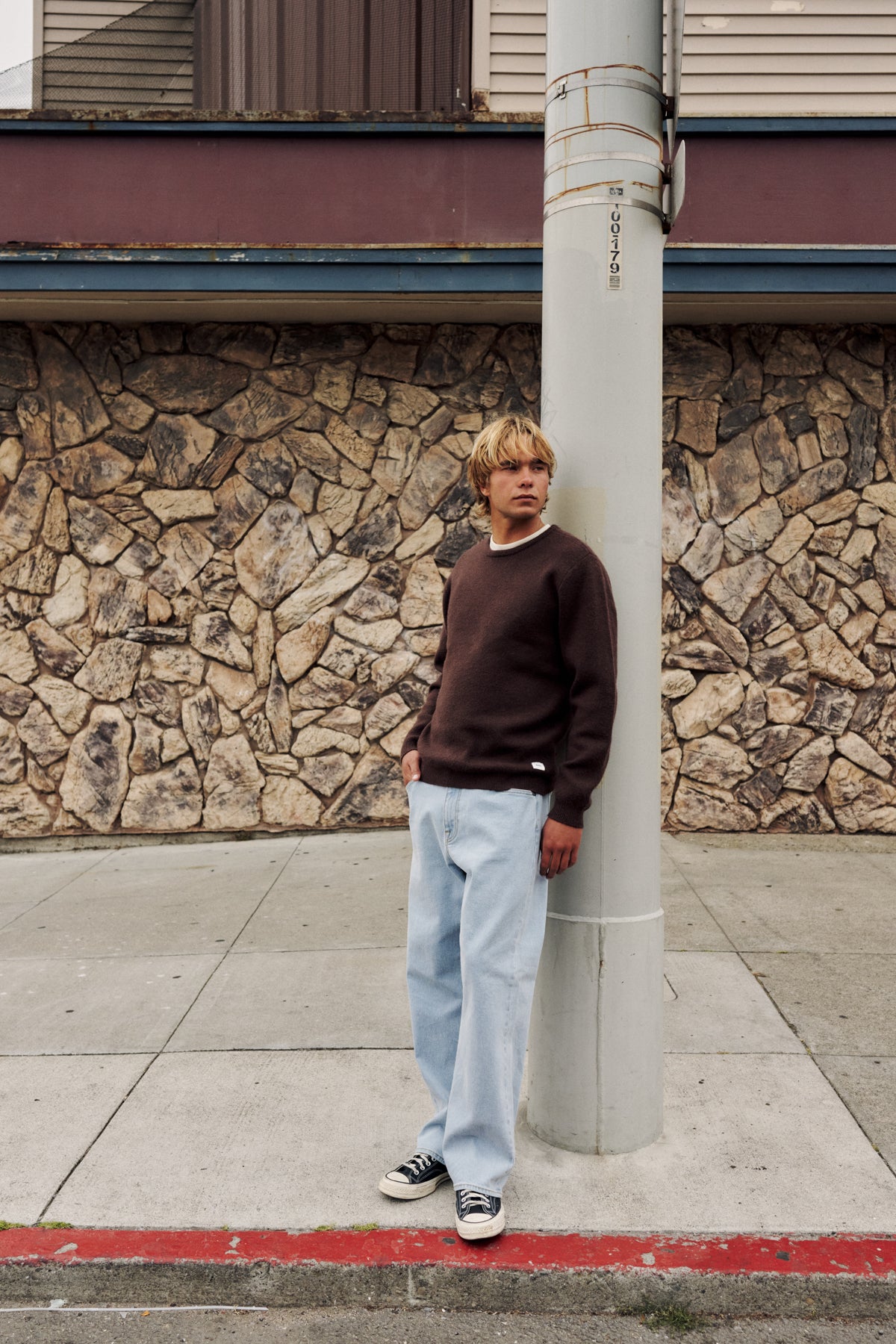 A young man with blonde hair stands leaning against a gray utility pole on a concrete sidewalk. He is wearing a dark brown crewneck sweatshirt, light blue baggy jeans, and black and white sneakers.