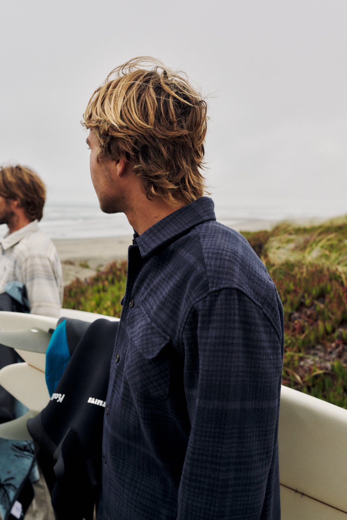 A man in a dark navy plaid button-down shirt stands on a beach dune with surfboards and the ocean in the background. Another person is partially visible in the blurred background.