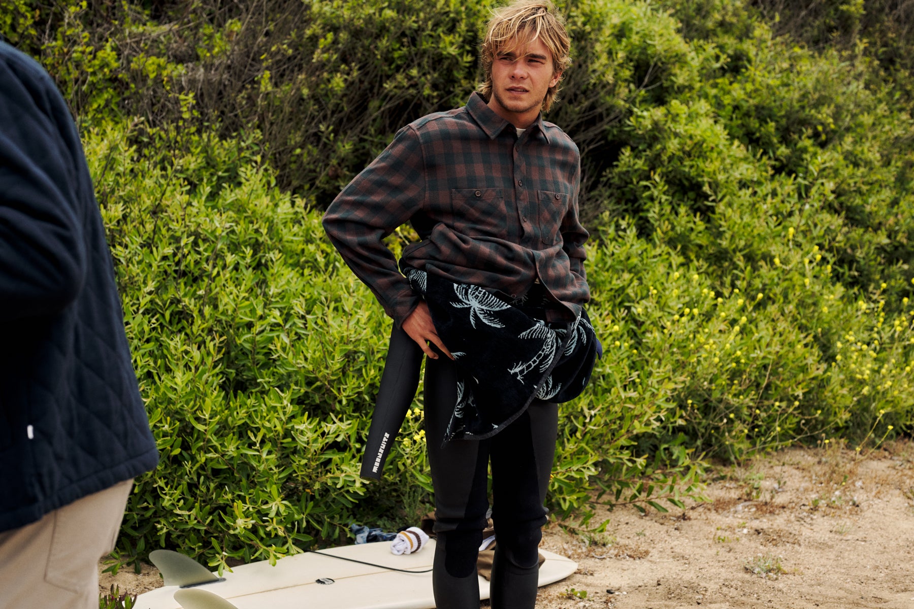 A man standing outdoors in front of dense green shrubs, wearing a dark plaid button-up shirt, black pants, and holding a black wetsuit or wrap with a white fish-like print. A surfboard is visible on the ground, and part of another person appears at the left edge.