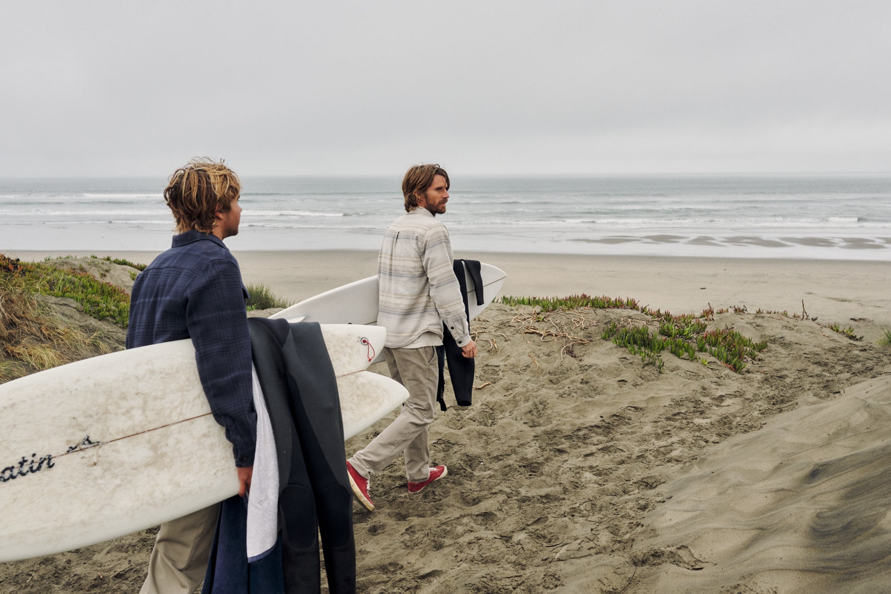 Two surfers walking along a sandy beach dune under an overcast sky, carrying surfboards and wetsuits. One surfer wears a navy blue jacket and beige pants, while the other wears a light gray plaid jacket, beige pants, and red sneakers.