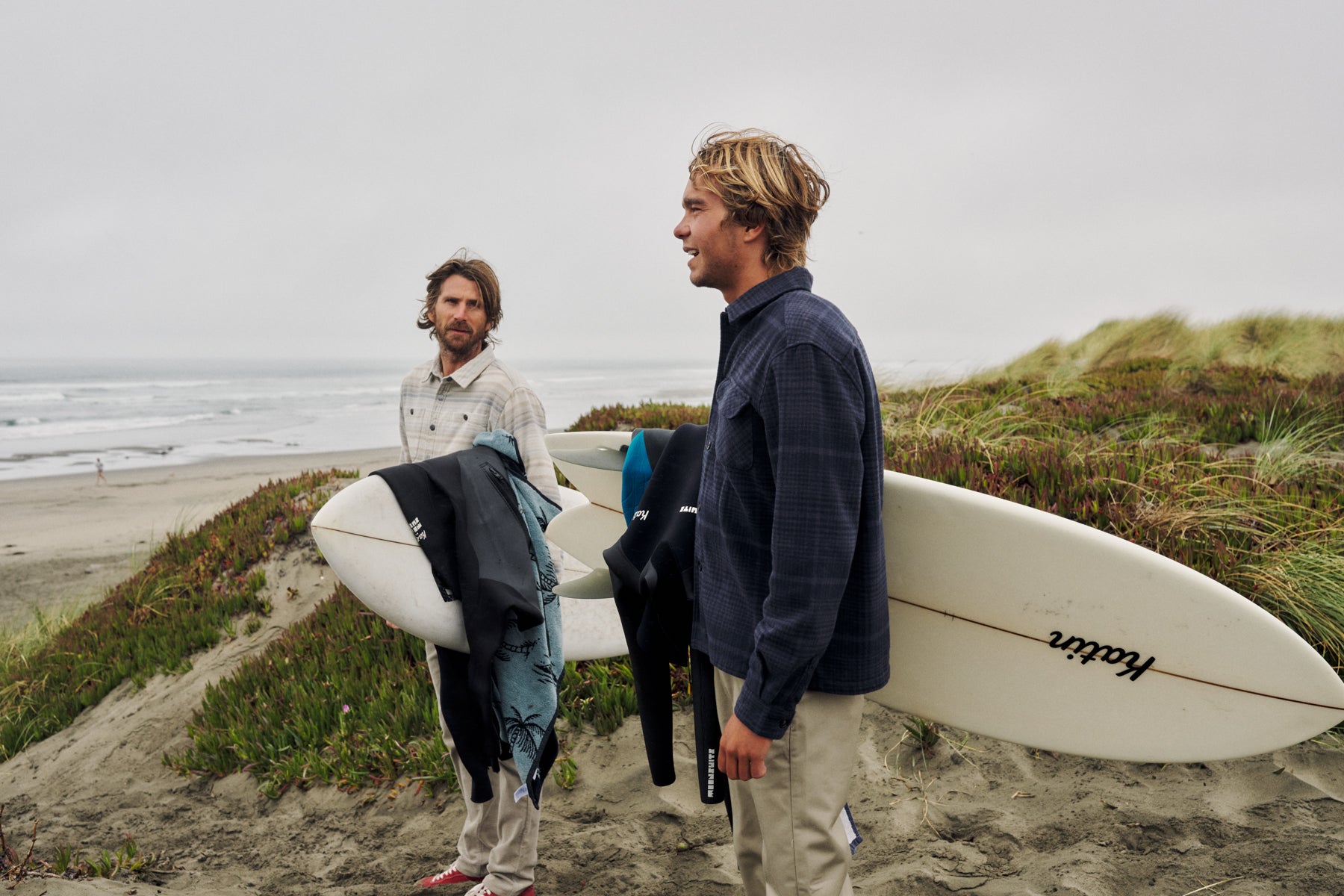 Two men standing on a sandy beach dune holding surfboards and wet suits, with the ocean and overcast sky in the background. One man wears a dark navy plaid shirt and khaki pants, while the other wears a light beige shirt.