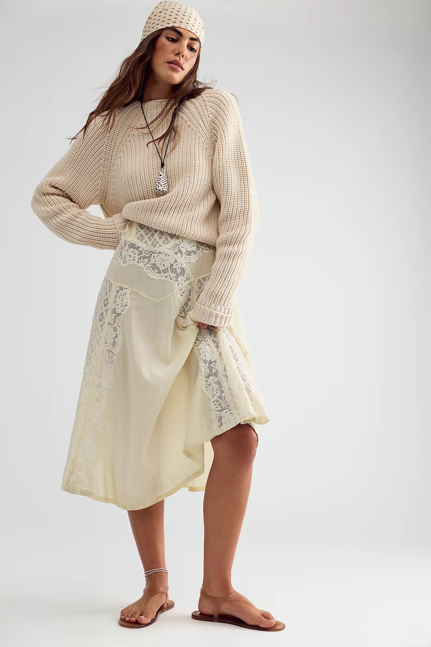 A woman modeling a cream-colored outfit consisting of a chunky knit sweater, a matching knit beanie, and a cream midi skirt with lace panels, standing against a light gray studio background.