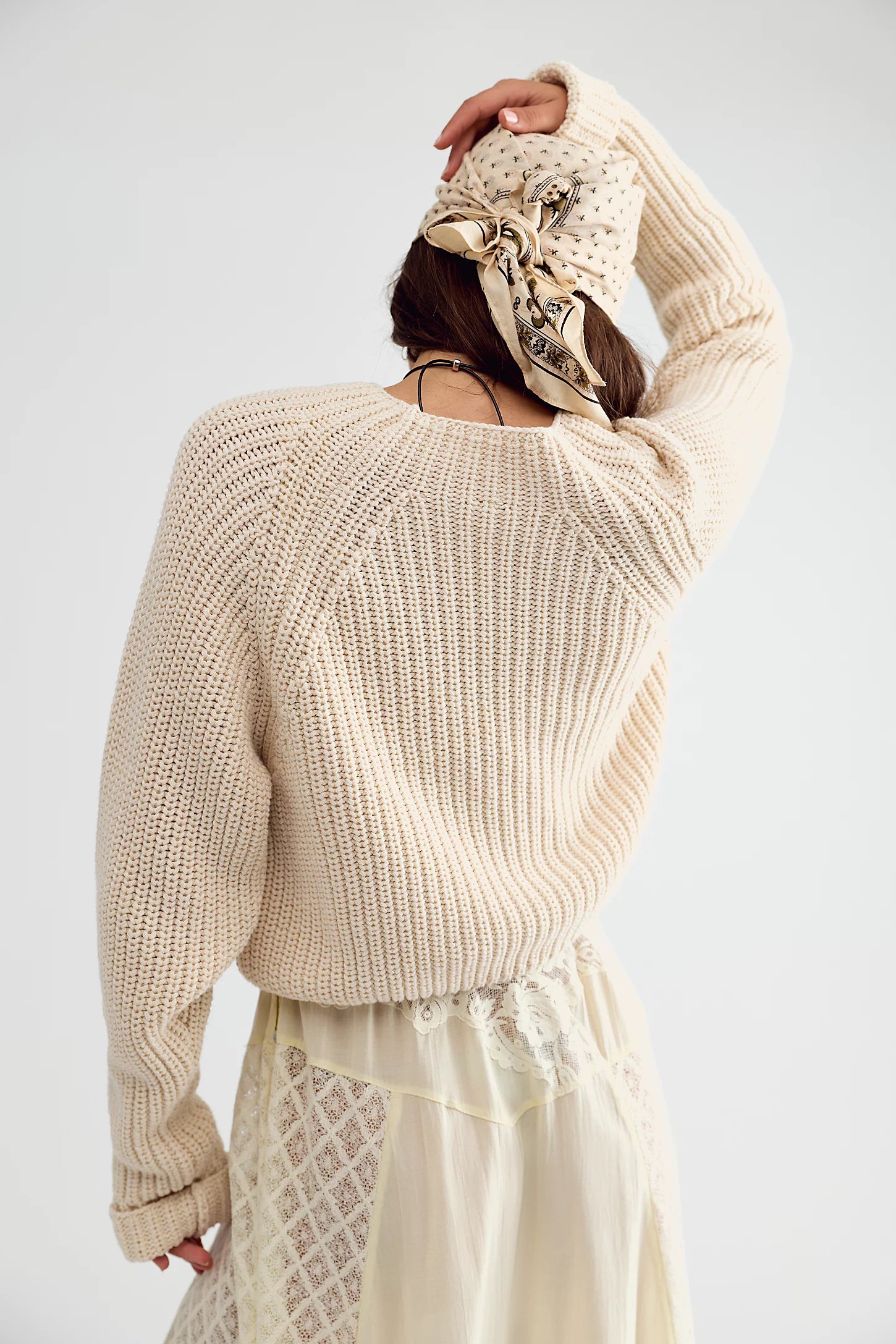 A woman shown from the back wearing a cream-colored, chunky ribbed knit sweater and a matching cream lace skirt, accessorized with a patterned beige headscarf, against a plain light gray studio background.