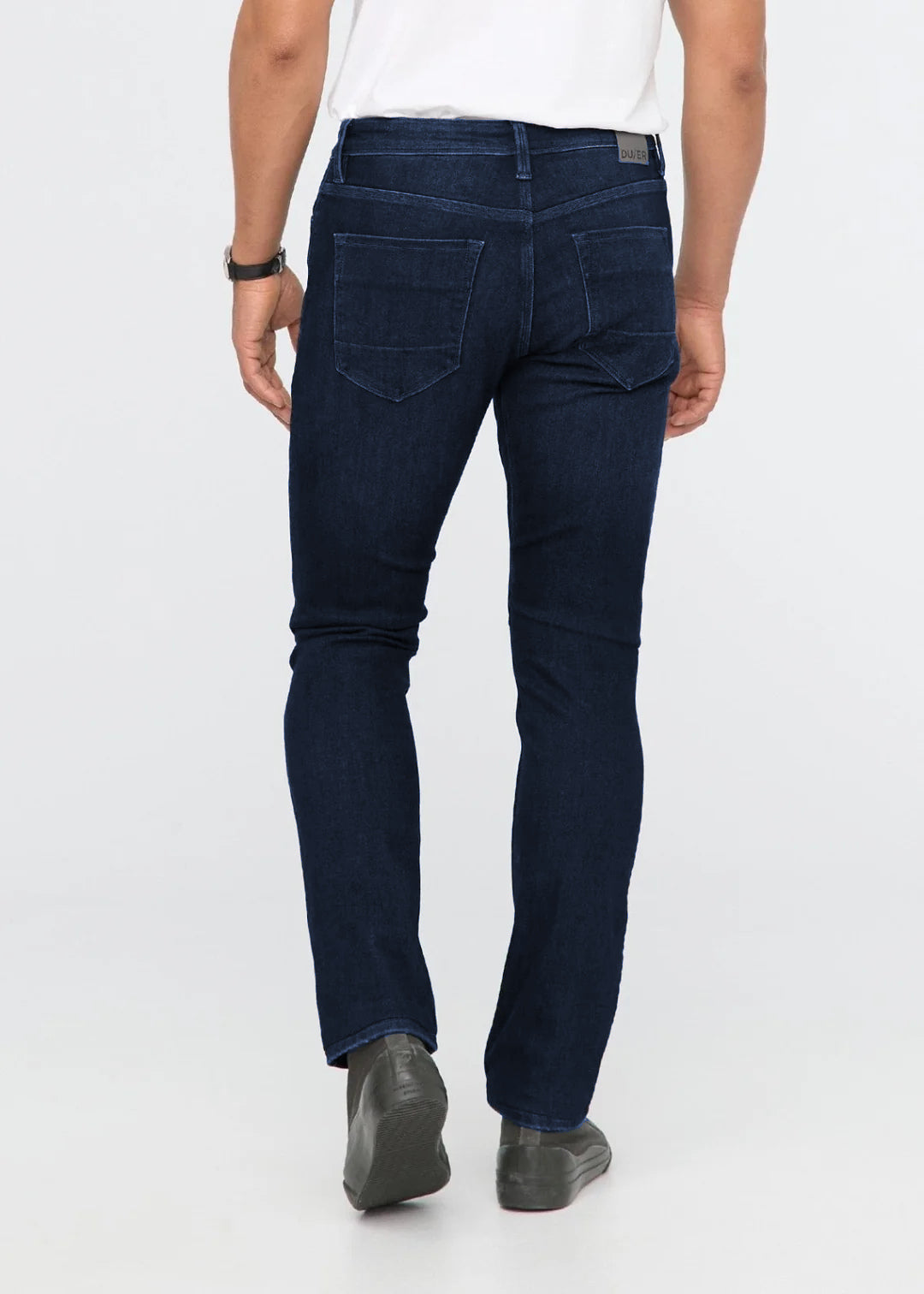 Rear view of a person wearing dark indigo slim-fit jeans with a white t-shirt and dark gray shoes, standing against a light gray studio background.