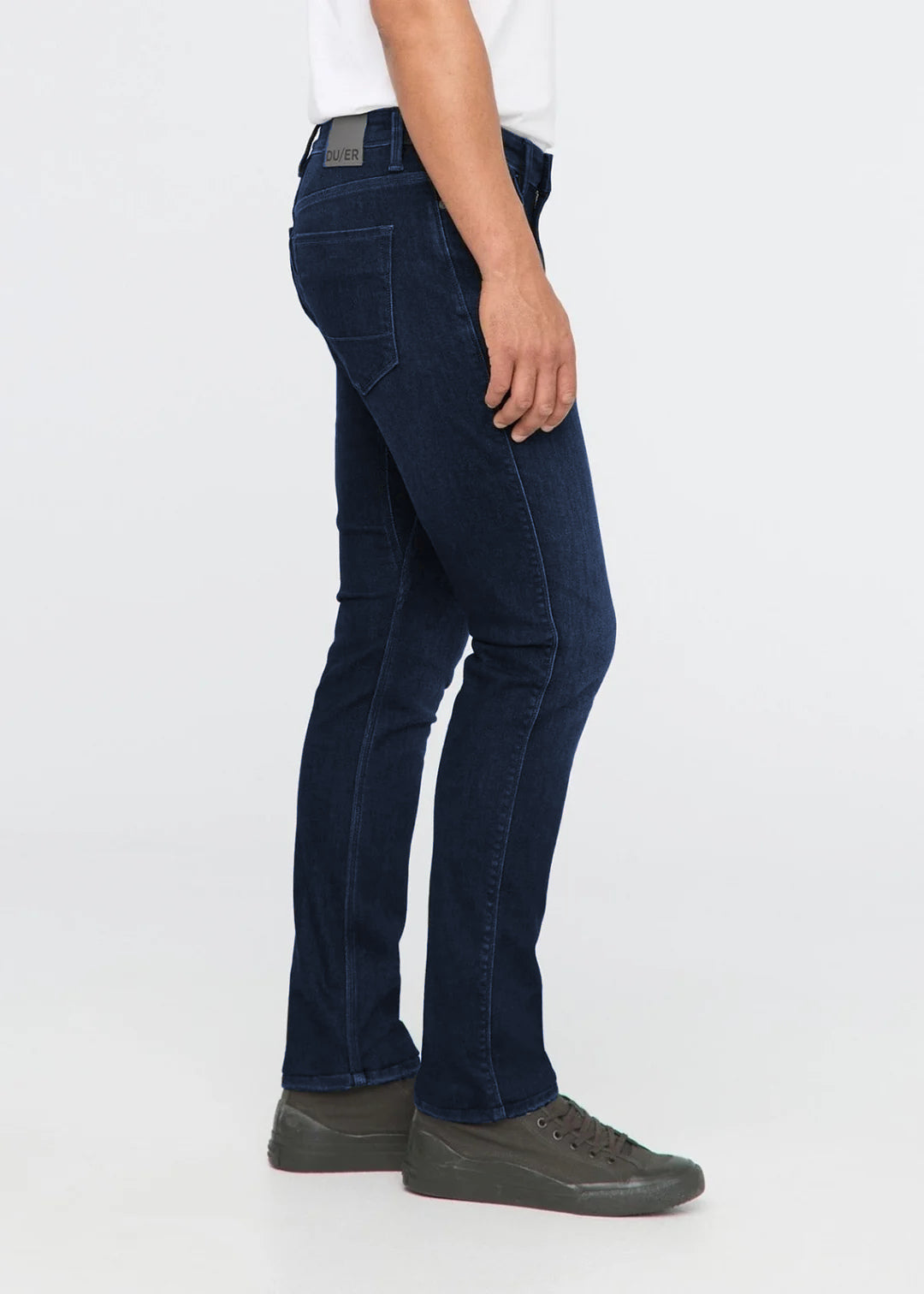 A side profile view of a person wearing dark navy blue slim-fit jeans, a white t-shirt, and dark gray sneakers against a plain white studio background.