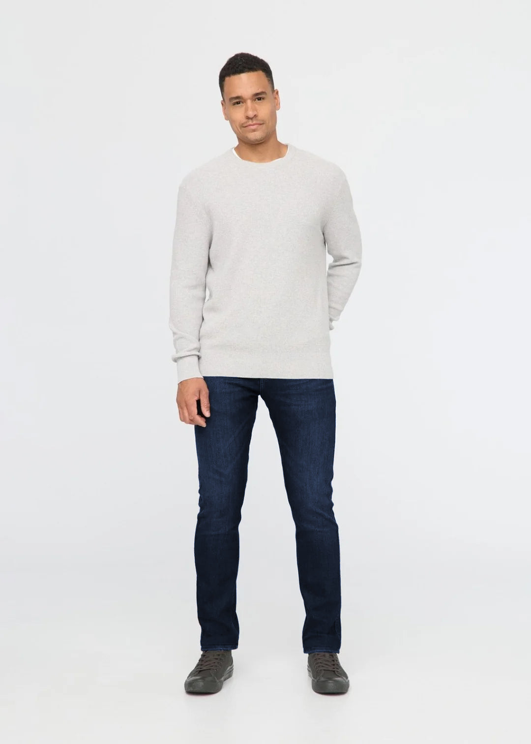 A man modeling a light gray crewneck sweater and dark blue jeans against a plain white studio background.