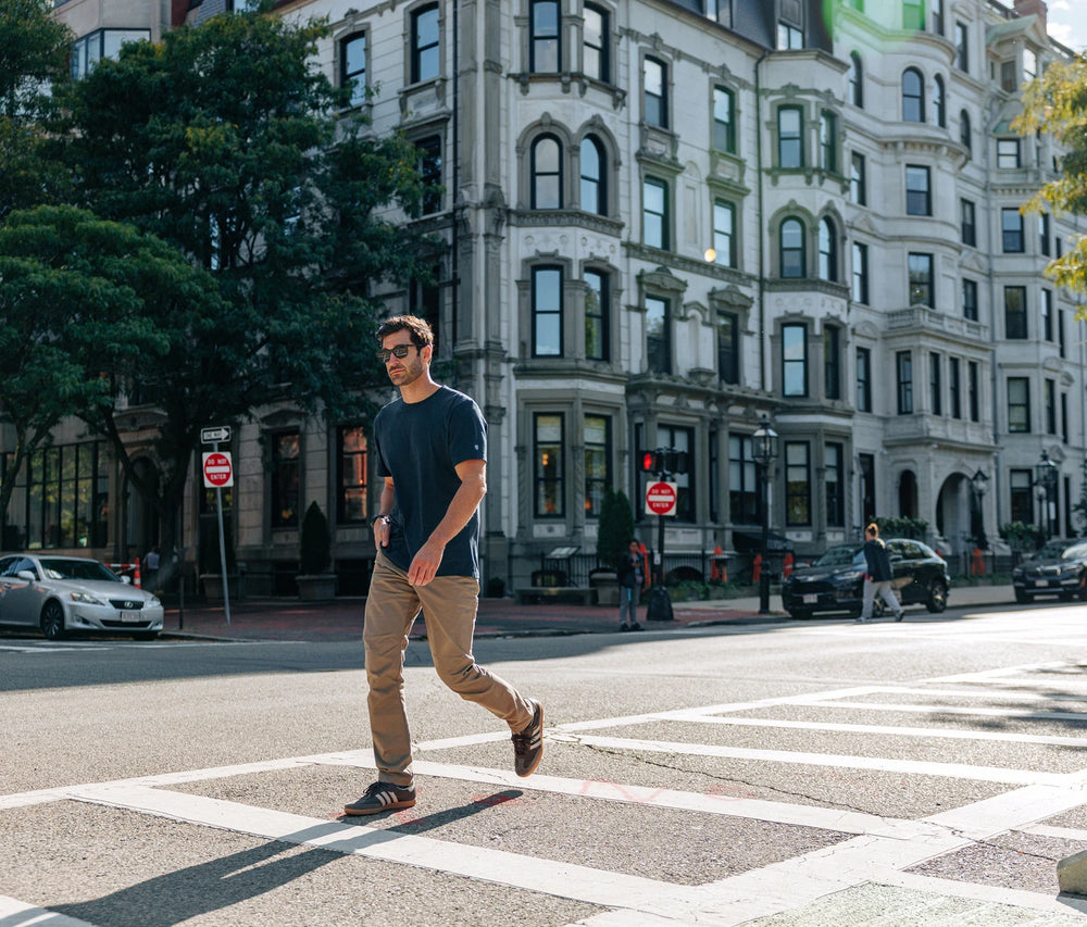 Man walking across a crosswalk in an urban setting with buildings and street signs in the background.