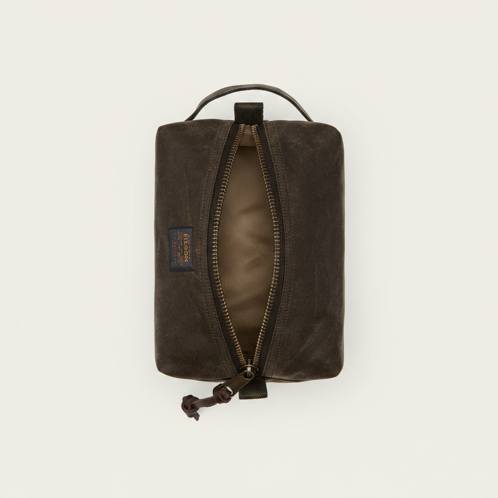 A dark brown waxed canvas zippered pouch with an open zipper showing a tan interior, featuring a top handle and a small brand patch on the side.